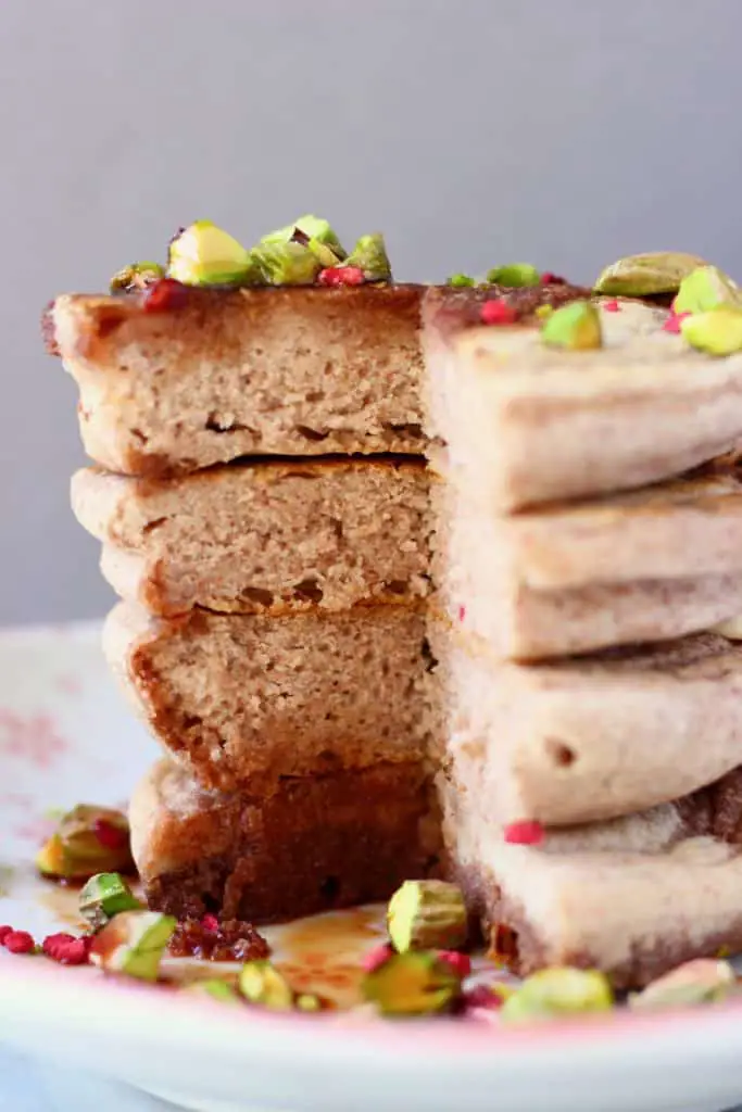 A stack of four buckwheat pancakes on a white plate decorated with chopped pistachios and freeze-dried raspberries covered in dark syrup against a grey background