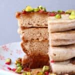 A stack of four buckwheat pancakes on a white plate decorated with chopped pistachios and freeze-dried raspberries covered in dark syrup against a grey background