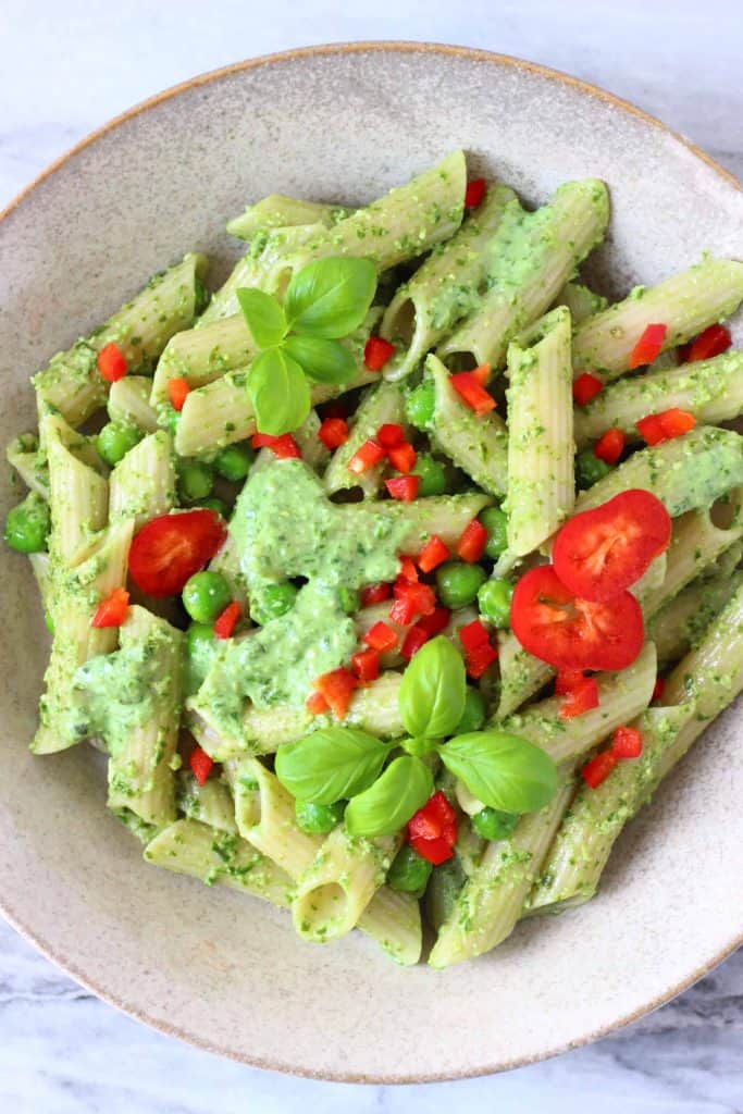 Penne pasta with pesto sauce, green peas and chopped red pepper in a beige bowl against a marble background