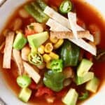 Tomato soup with white beans, sweetcorn and green peppers topped with tortilla strips and diced avocado in a white bowl against a white background