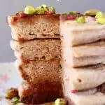 A stack of four buckwheat pancakes on a white plate decorated with chopped pistachios and freeze-dried raspberries covered in dark syrup against a grey background