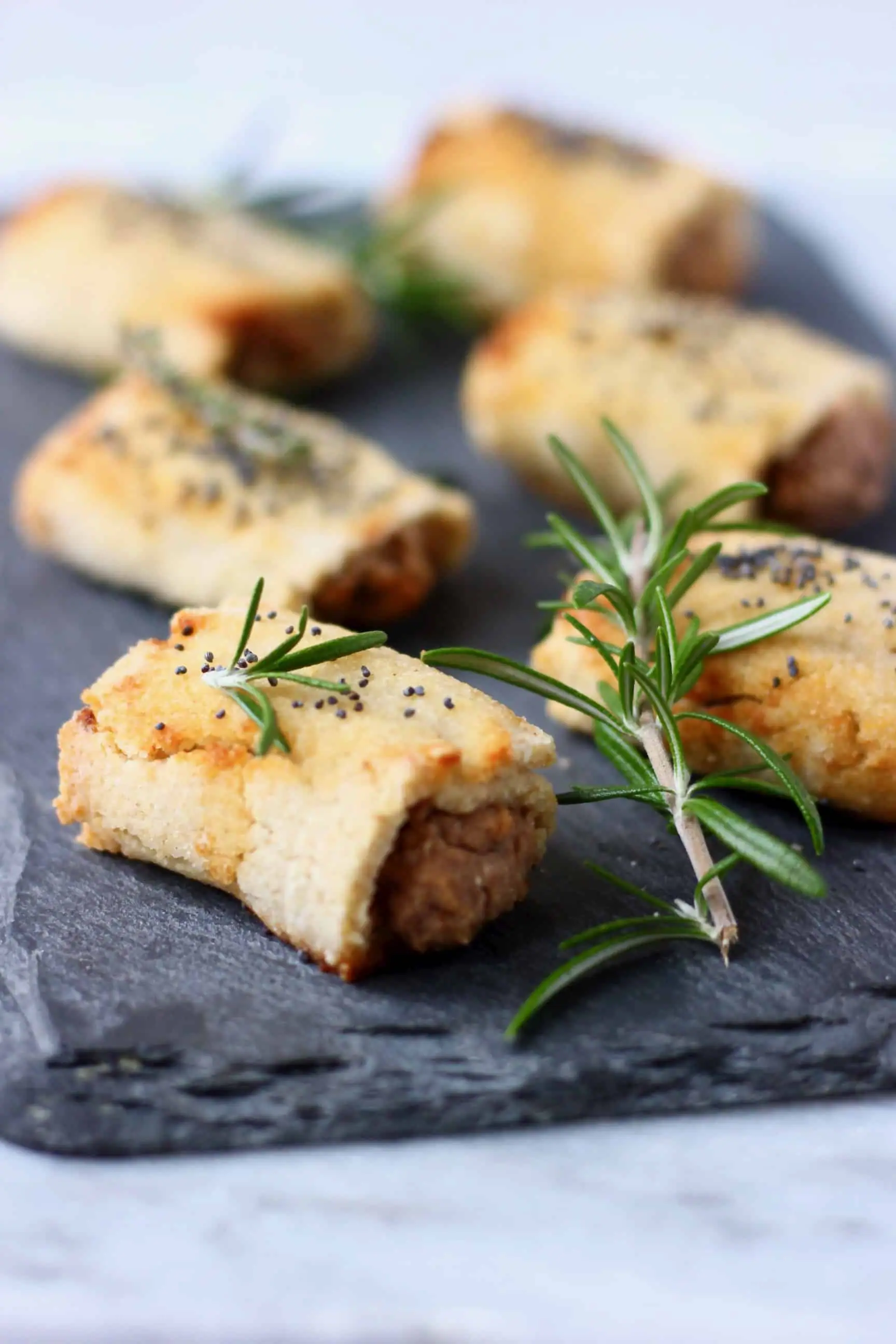 Six vegan sausage rolls sprinkled with poppy seeds on a black slab decorated with sprigs of rosemary