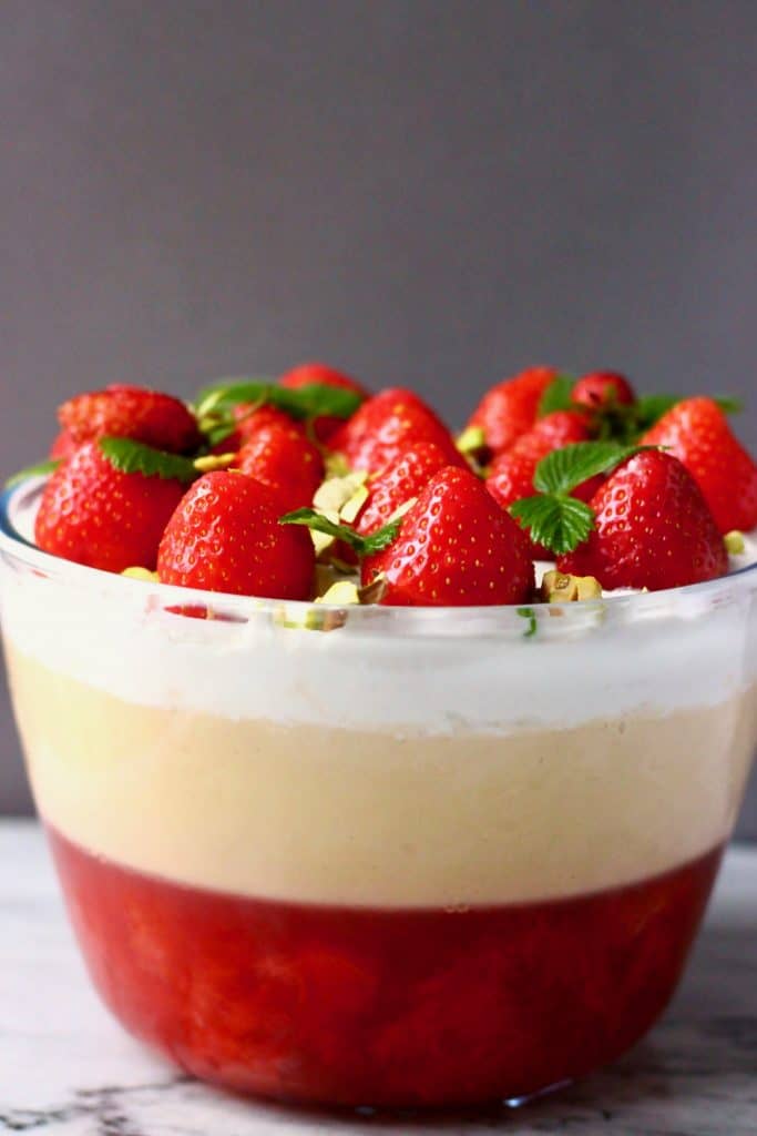 Photo of red strawberry jelly topped with yellow custard, white cream, fresh strawberries, green leaves and chopped pistachio nuts against a grey background