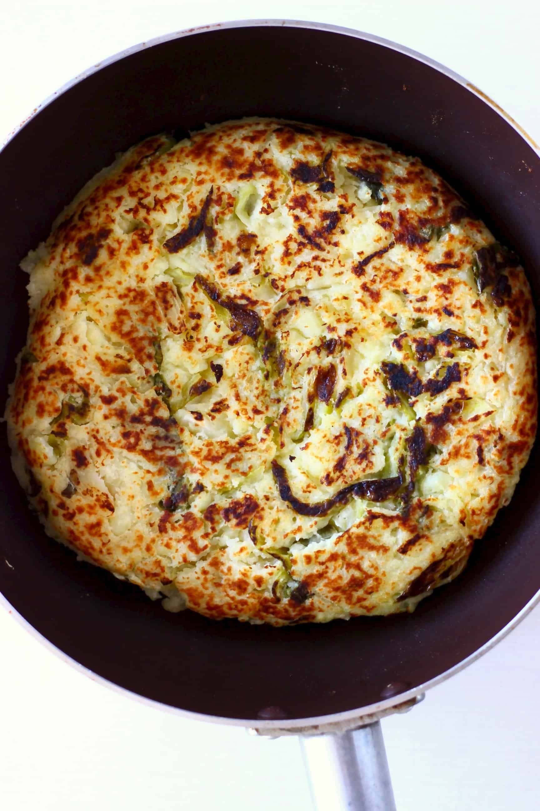 A circular, golden brown bubble and squeak cake in a dark frying pan against a white background