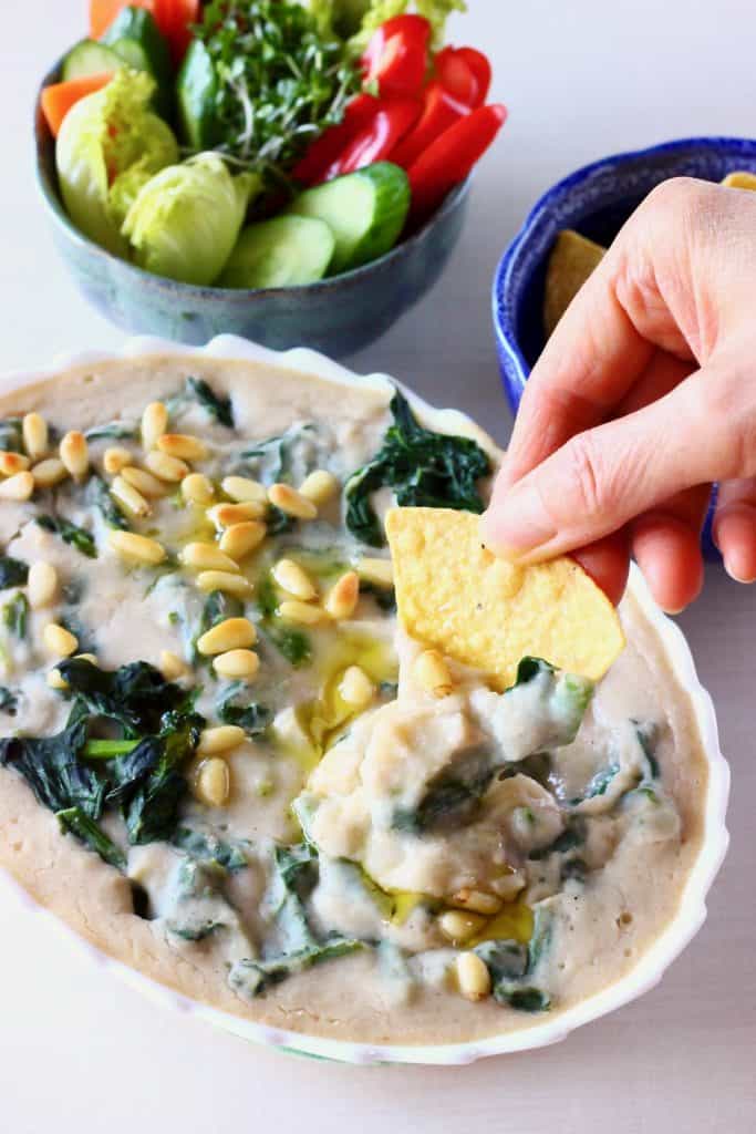 Photo of an oval dish filled with a white dip with chopped artichokes and spinach topped with pine nuts with a hand dipping a tortilla chip into it