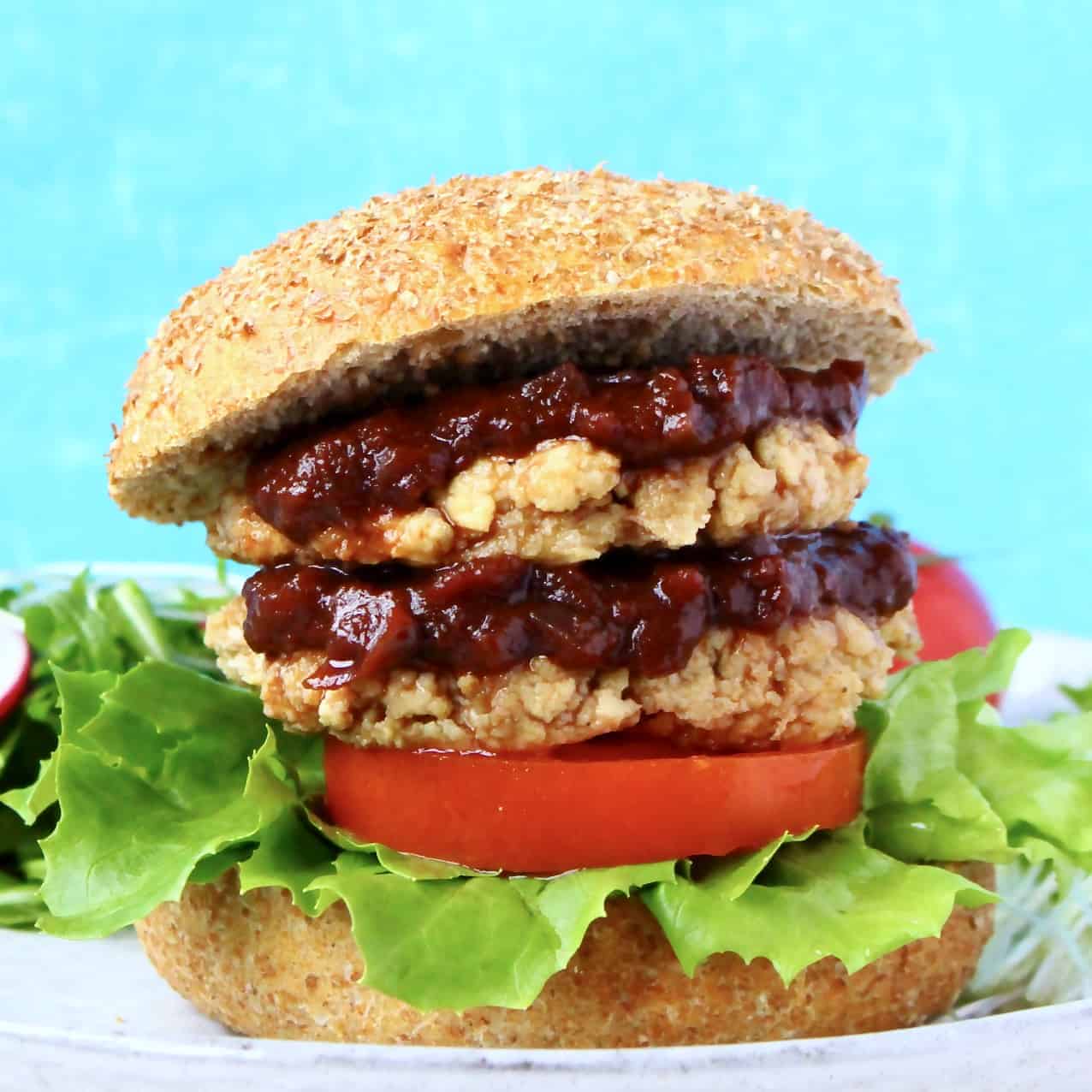 A tofu burger with two patties, brown sauce, lettuce and tomato in a burger bun on a grey plate against a blue background