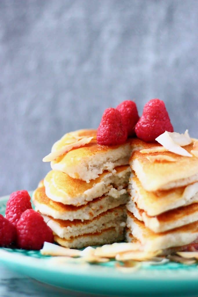 Five coconut flour pancakes stacked in a pile decorated with fresh raspberries and coconut flakes on a green plate against a grey background