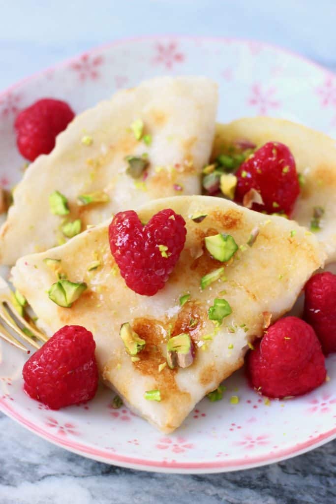 Three folded crepes on a white plate with pink flowers decorated with fresh raspberries and chopped pistachios with a gold fork against a marble background