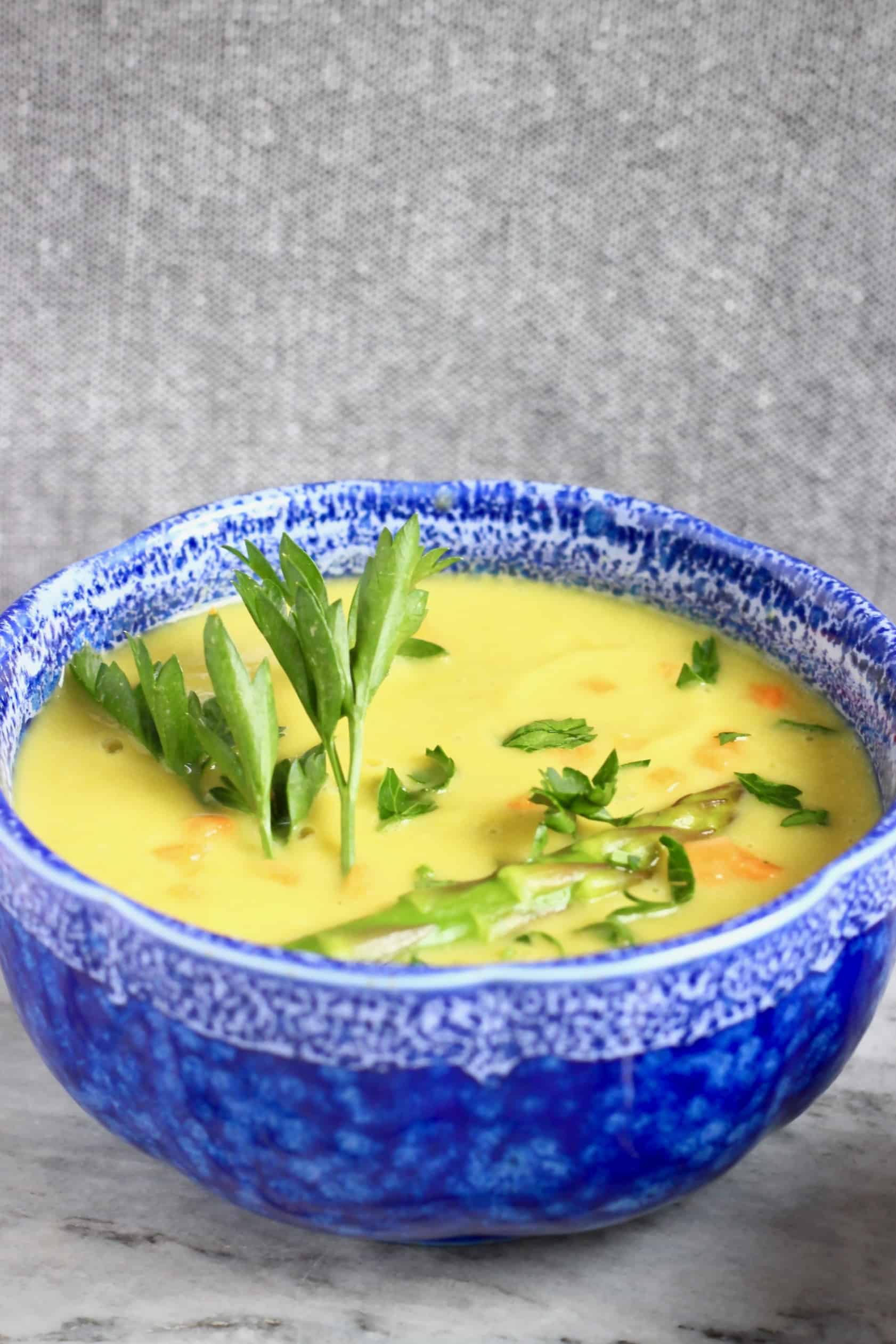 A dark blue bowl filled with light green asparagus soup decorated with green herbs against a grey background