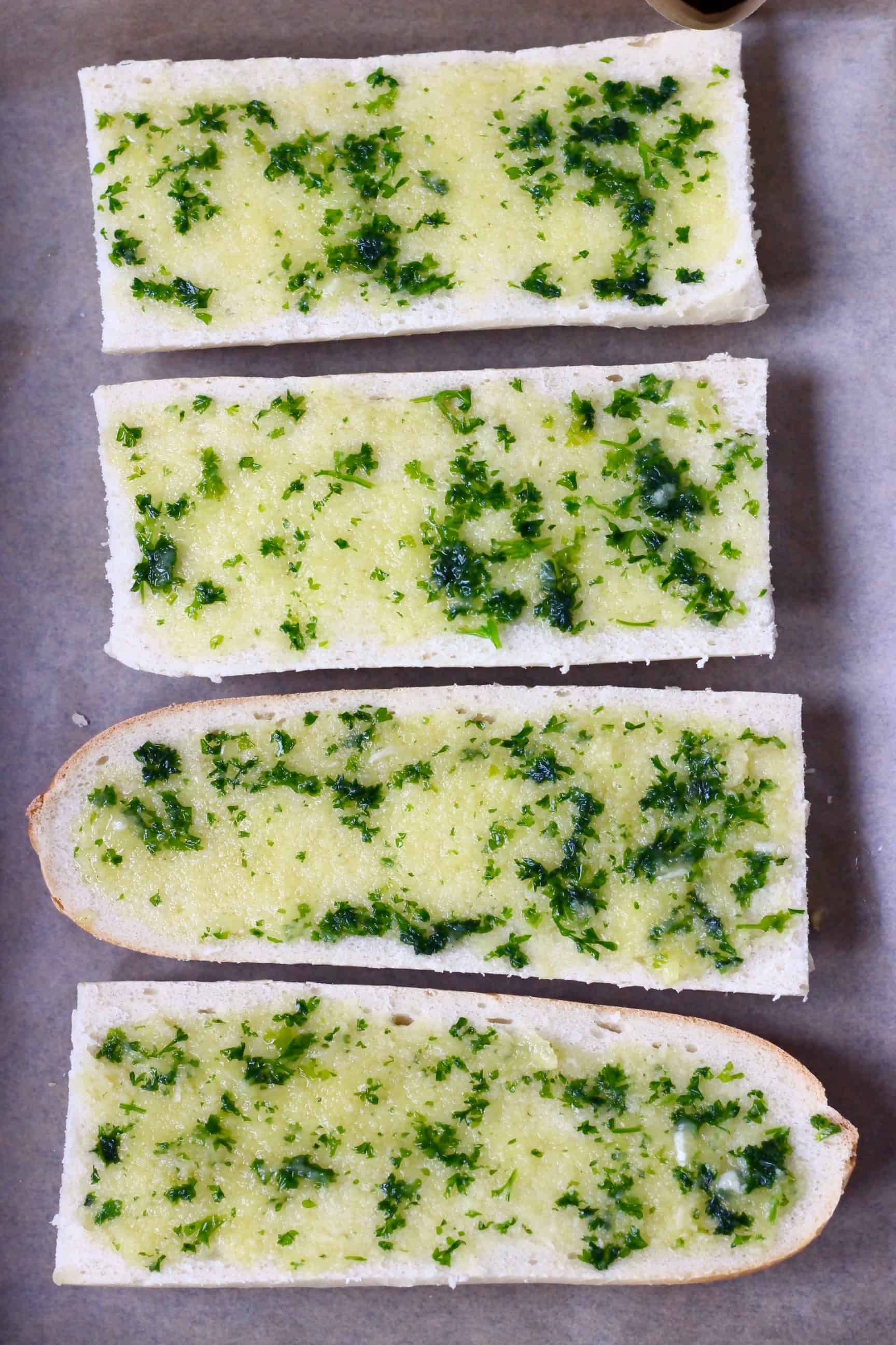 Four pieces of sliced white baguette spread with garlic and herb oil on a sheet of baking paper