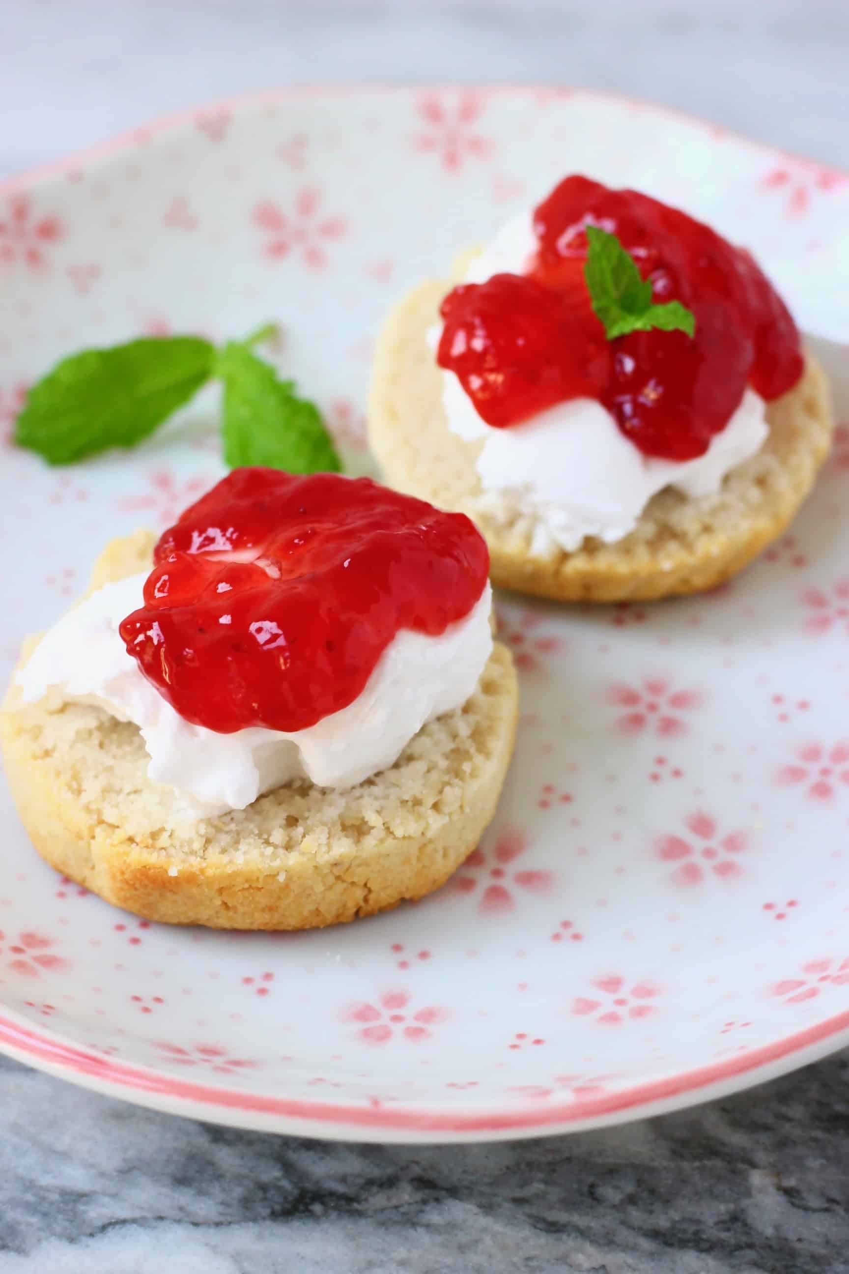 Two gluten-free vegan scone halves topped with white cream and strawberry jam decorated with mint leaves on a plate