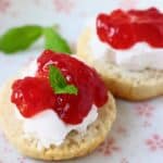 Two gluten-free vegan scones halves topped with white cream and strawberry jam decorate with mint leaves on a white plate with pink flowers
