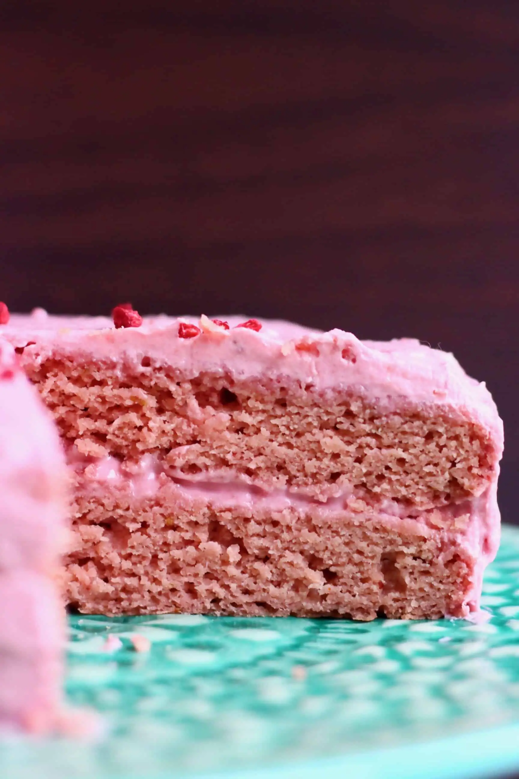 A sliced pink sponge cake with pink frosting on a green cake stand against a dark brown background