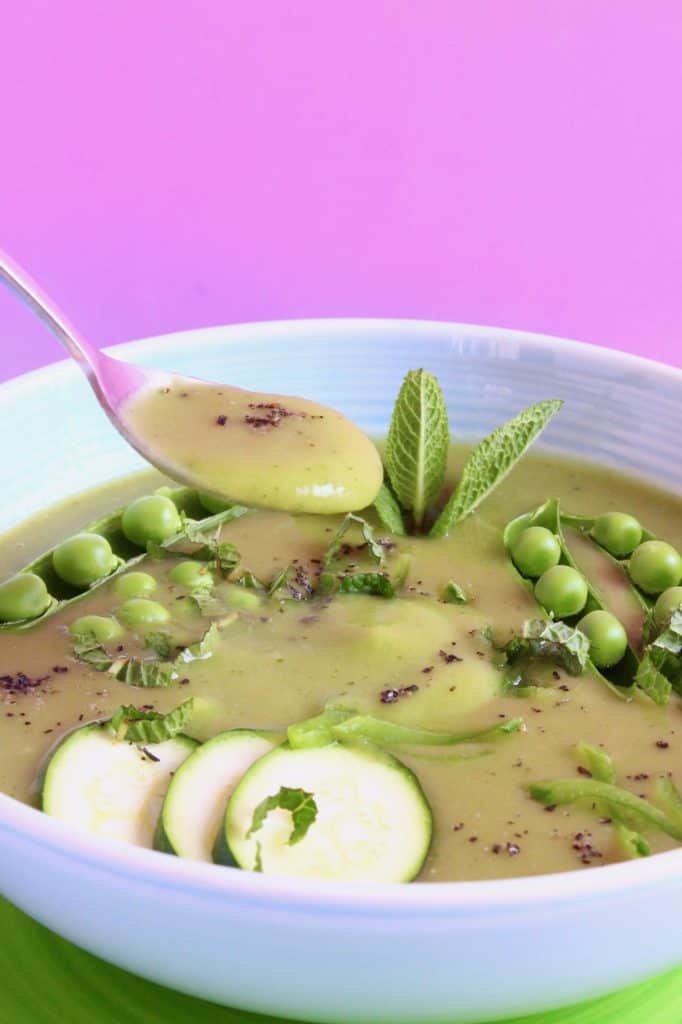Green soup topped with zucchini slices and peas in a blue bowl against a pink background