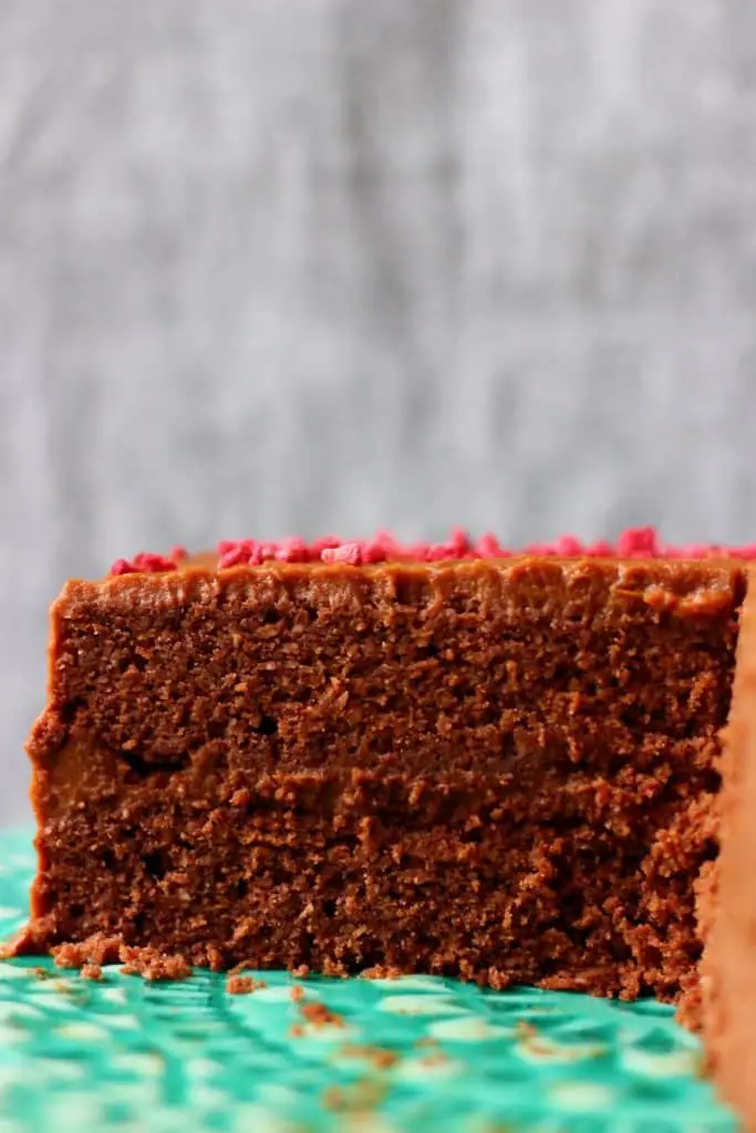 A sliced chocolate cake sandwiched with chocolate frosting and covered in chocolate frosting sprinkled with pink freeze-dried raspberries on a green cake stand against a grey background