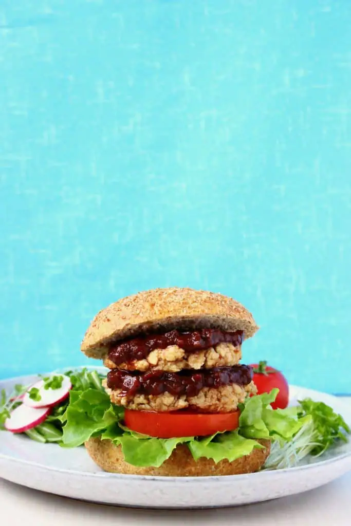A burger with two tofu patties, brown sauce, lettuce and tomato in a burger bun on a grey plate against a blue background