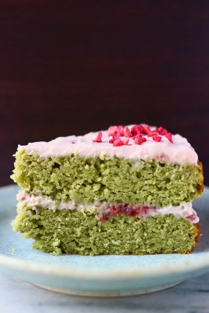 A slice of matcha sponge cake with strawberry frosting on a blue plate against a dark brown background