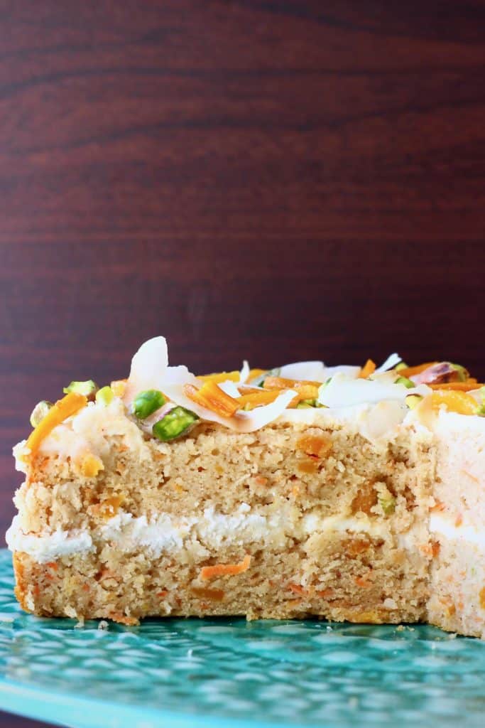 A carrot cake sponge cake with white frosting decorated with dried mango, chopped pistachios and coconut flakes on a green cake stand against a dark brown background