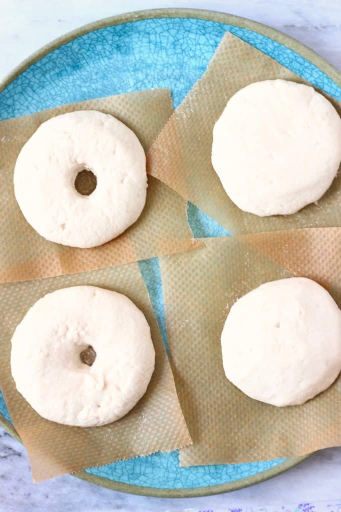 Four raw bagels on pieces of brown baking paper on a blue plate against a marble background