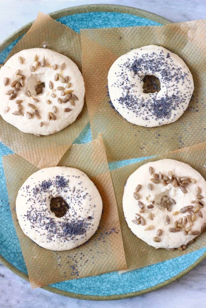 Four raw bagels topped with seeds on pieces of brown baking paper on a blue plate against a marble background