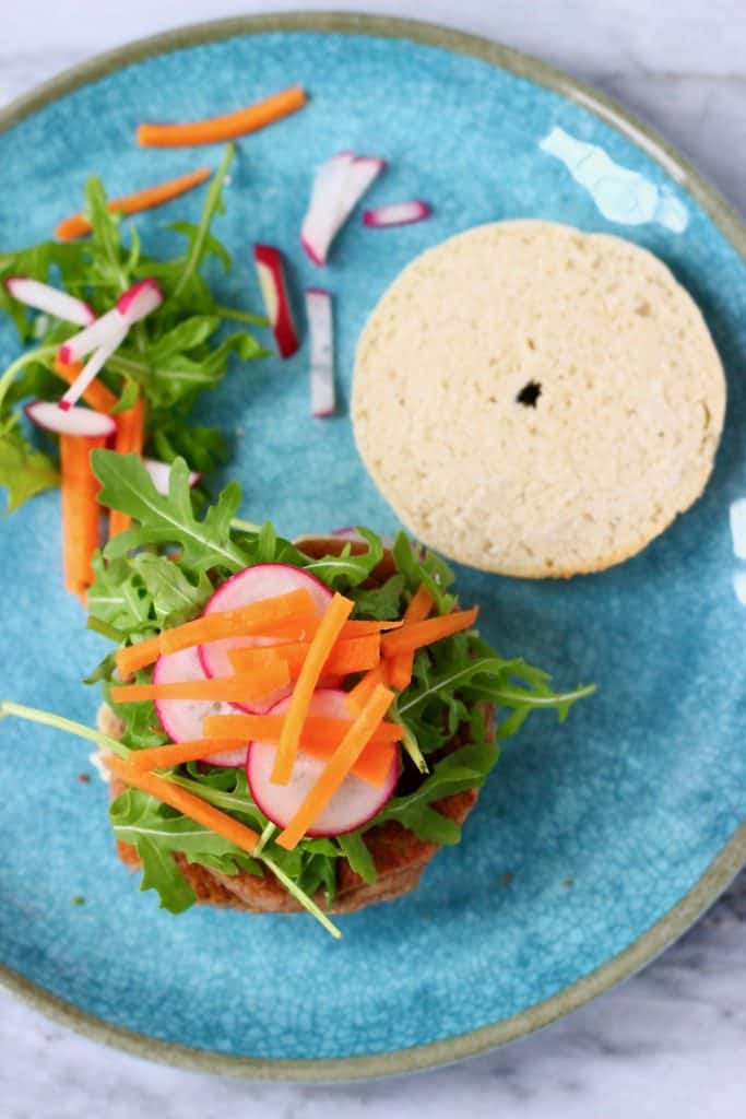 Half a bagel topped with vegan sausage and salad on a blue plate against a marble background