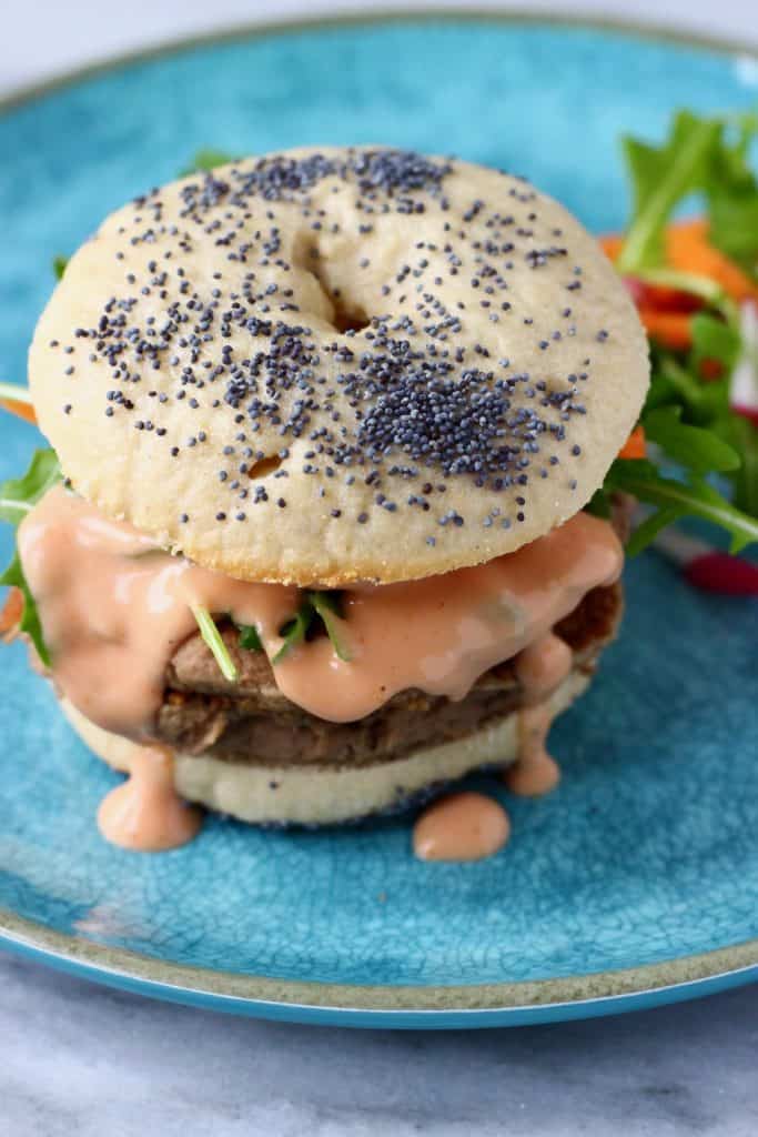 A bagel burger with vegan sausage patties, pink sauce and salad on a blue plate