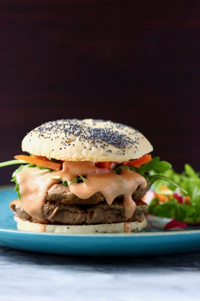 A bagel burger with vegan sausage patties, pink sauce and salad on a blue plate against a dark brown background