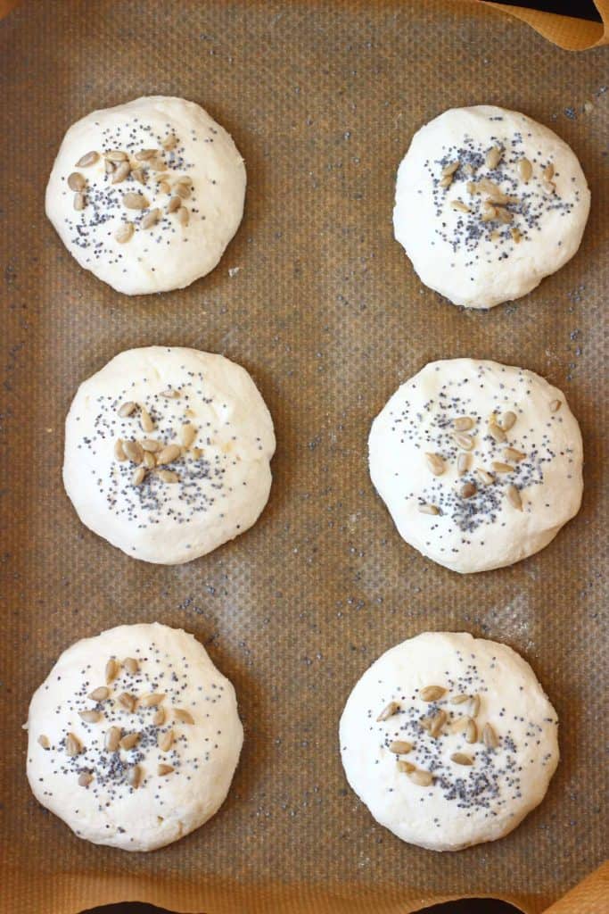 Six raw bread rolls topped with seeds on a sheet of brown baking paper