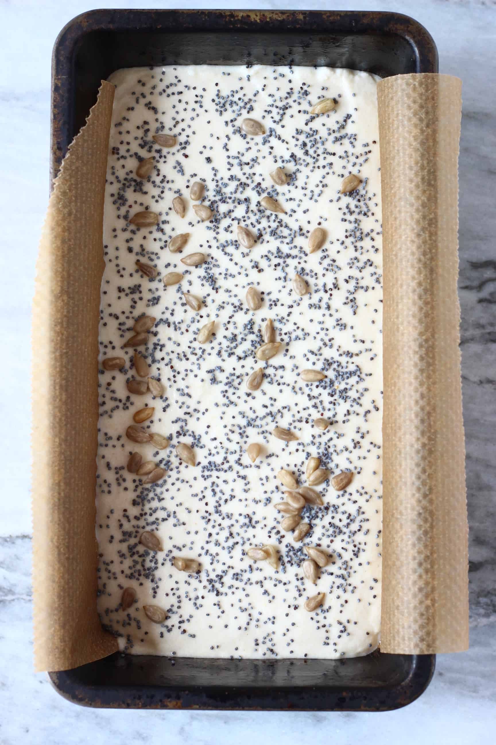 White gluten-free rice bread batter topped with seeds in a black loaf tin lined with baking paper 