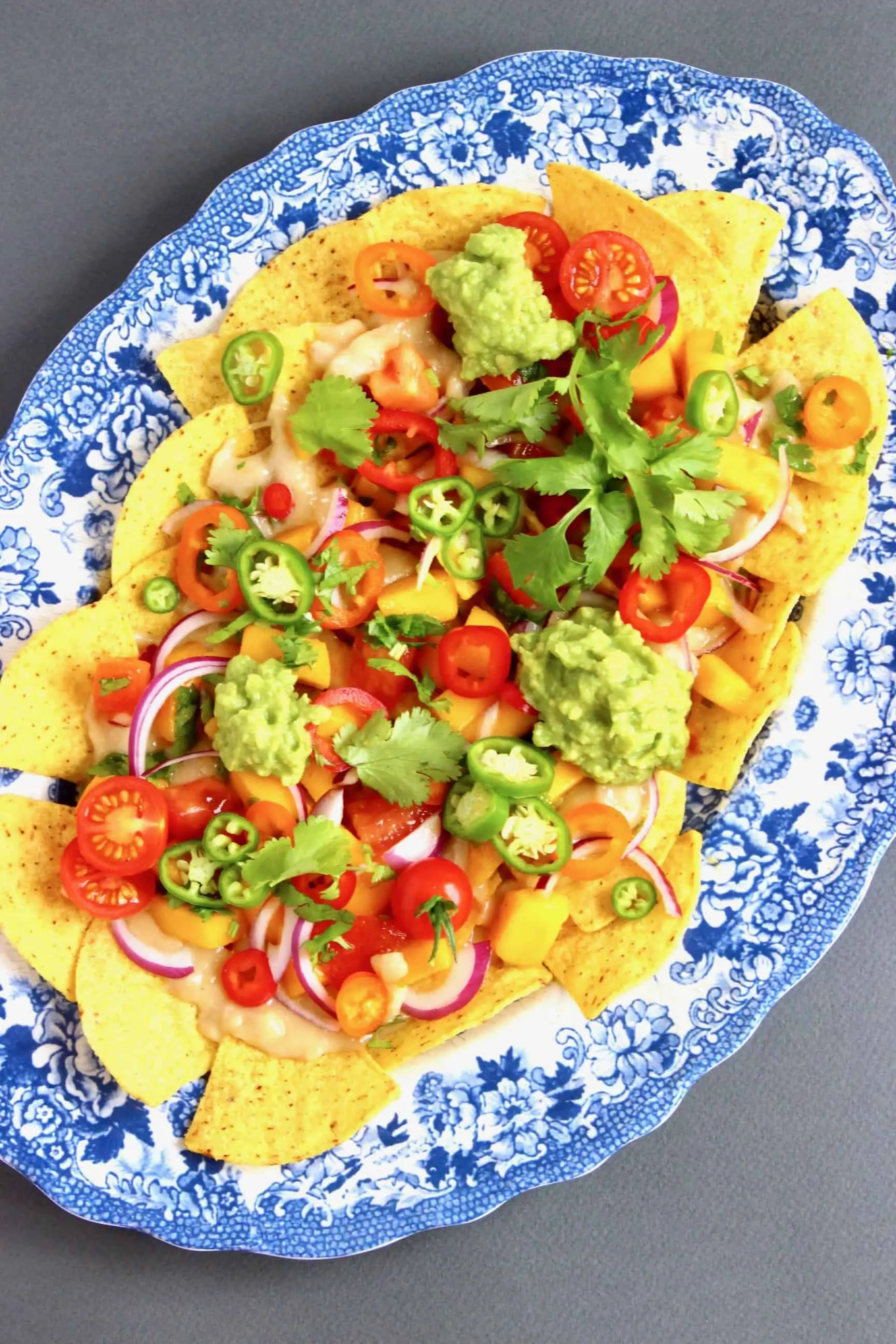 Nachos topped with lots of different vegetables on a oval blue plate against a grey background