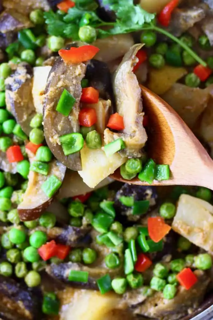 Aubergine, potato and pea curry in a pan with a wooden spoon lifting up a mouthful