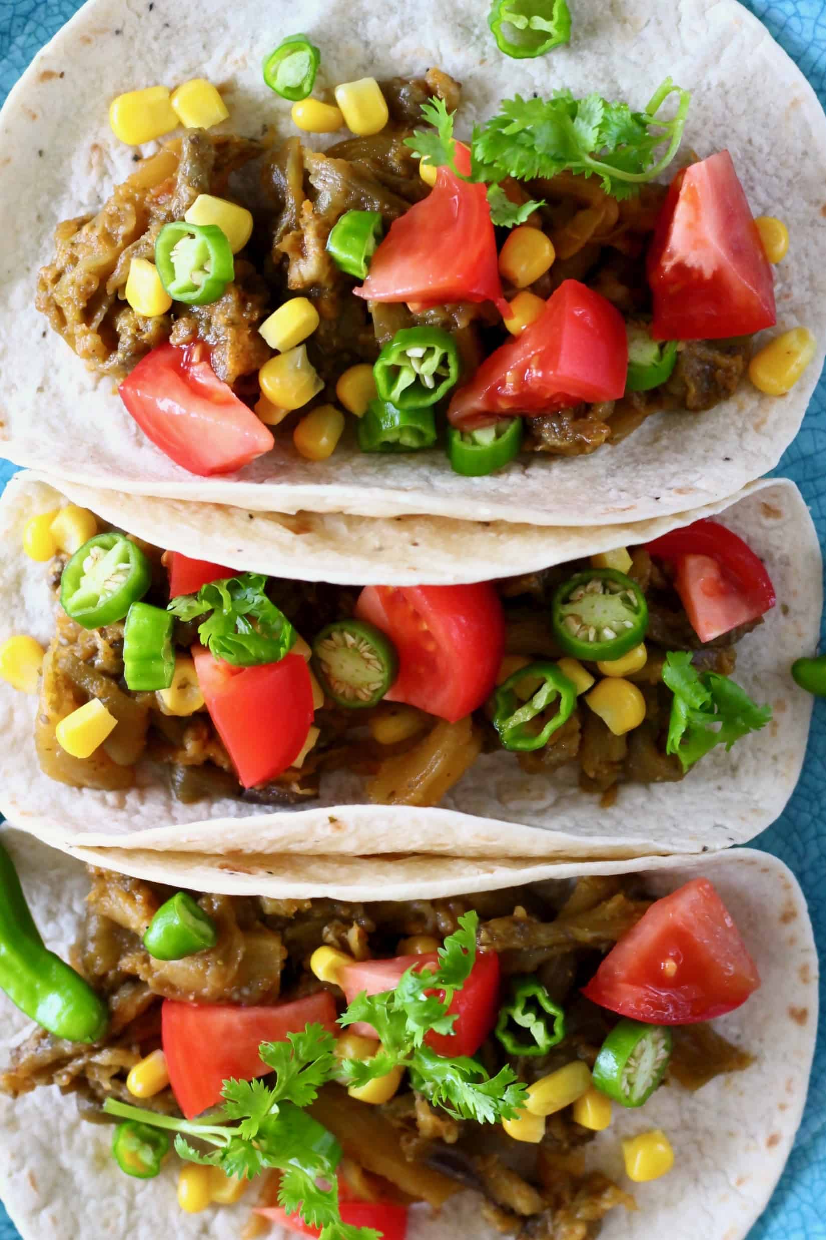 Photo of three tacos on a blue plate filled with roasted eggplant, sweetcorn, sliced green chilli, chopped tomatoes