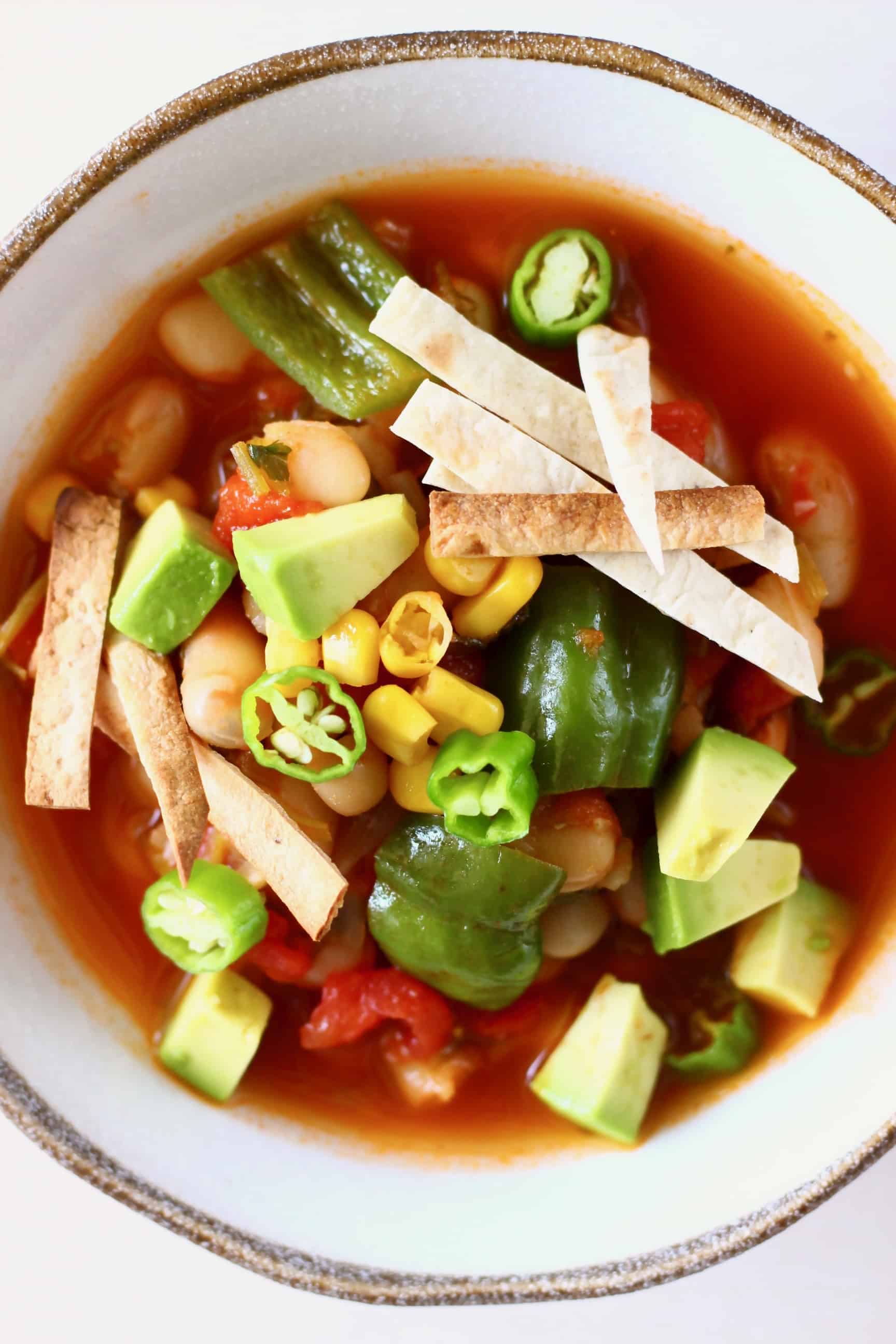 Tomato soup with white beans, sweetcorn and green peppers topped with tortilla strips and diced avocado in a white bowl against a white background