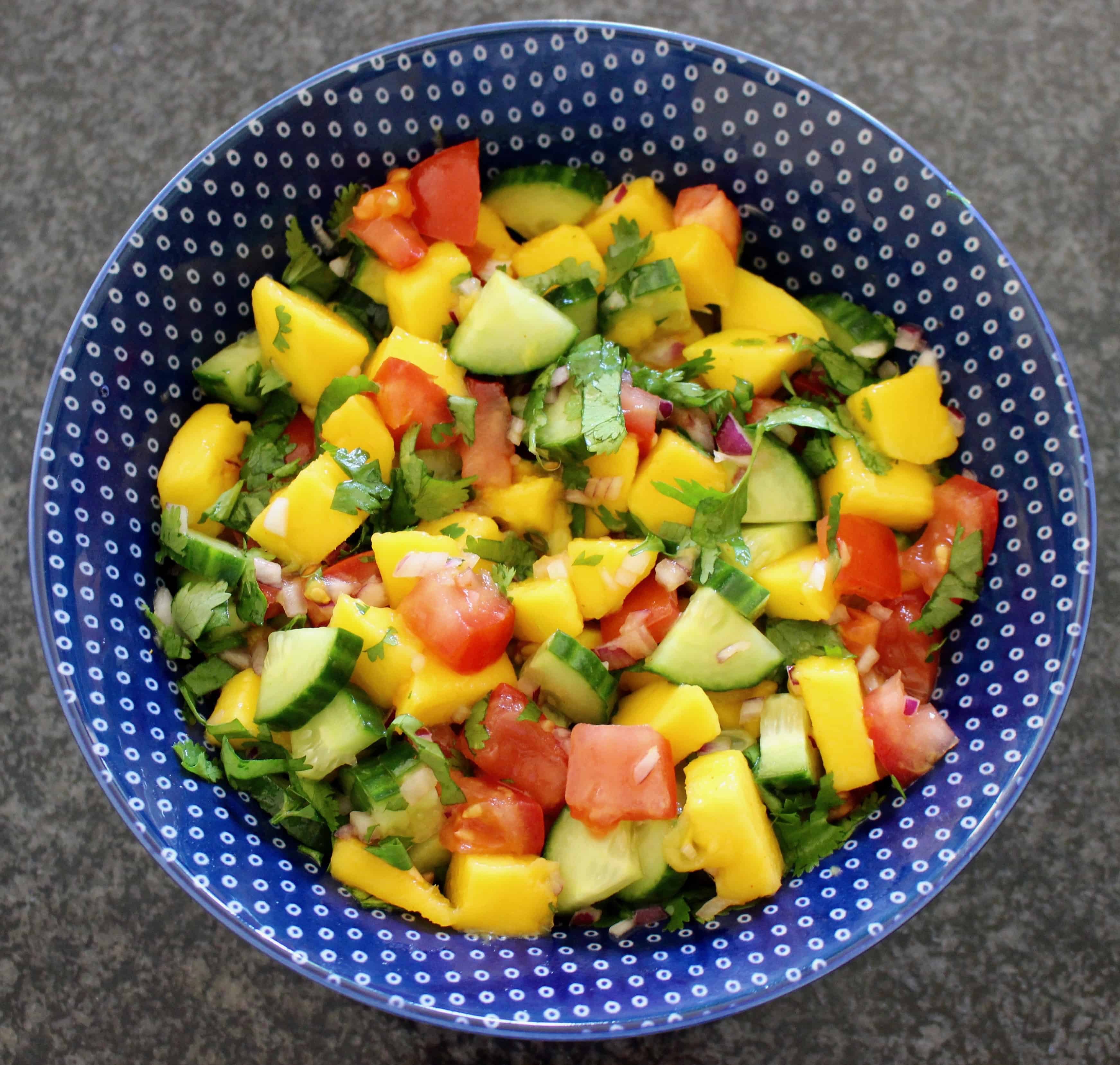 Chopped mango, tomatoes, cucumber and coriander in a blue bowl against a granite background