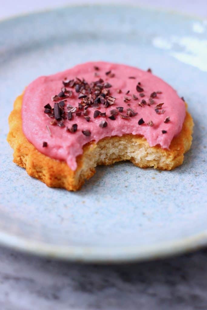 Photo of a circular cookie topped with pink frosting and brown cacao nibs with a bite taken out of it on a blue plate
