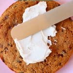 Photo of a fruit cake sponge on a pink plate with a silver palette knife spreading white frosting on top of it