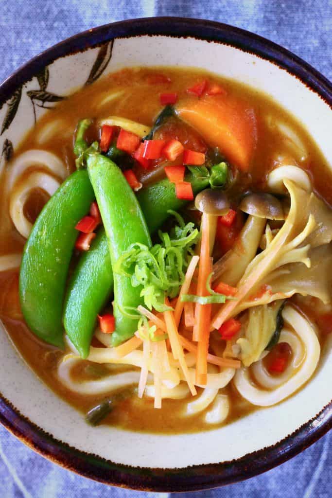 Udon noodles and vegetables in a brown curry sauce in a white bowl with a dark brown rim against a blue background