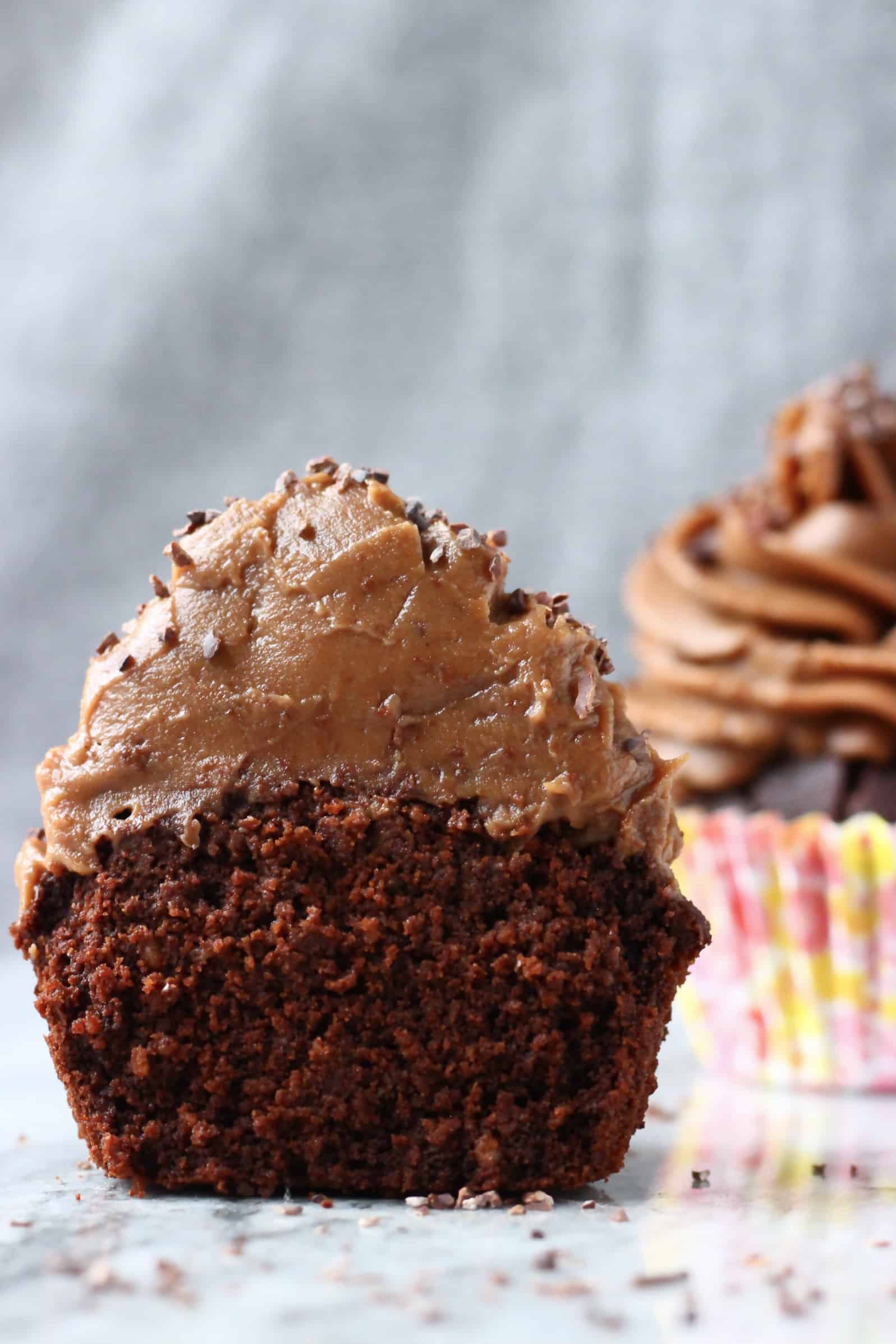 A gluten-free vegan chocolate cupcake with frosting cut in half with a cupcake in the background