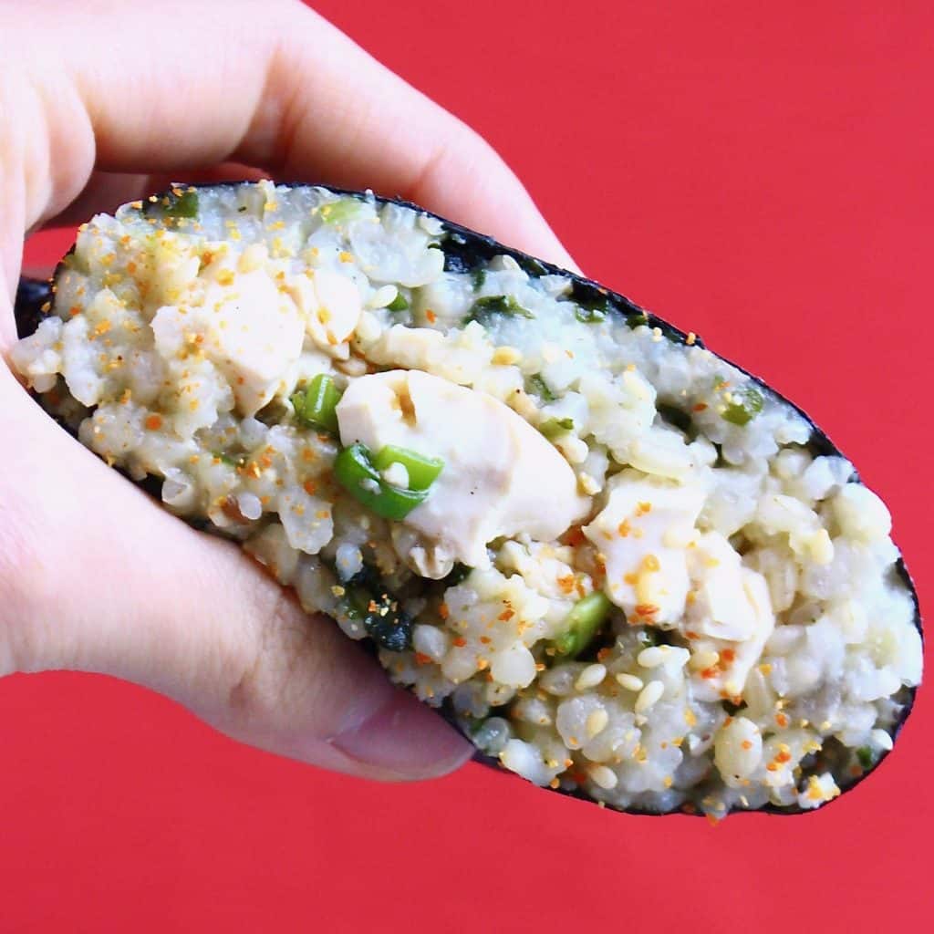 A sushi sandwich filled with scrambled tofu being held up by a hand against a red background
