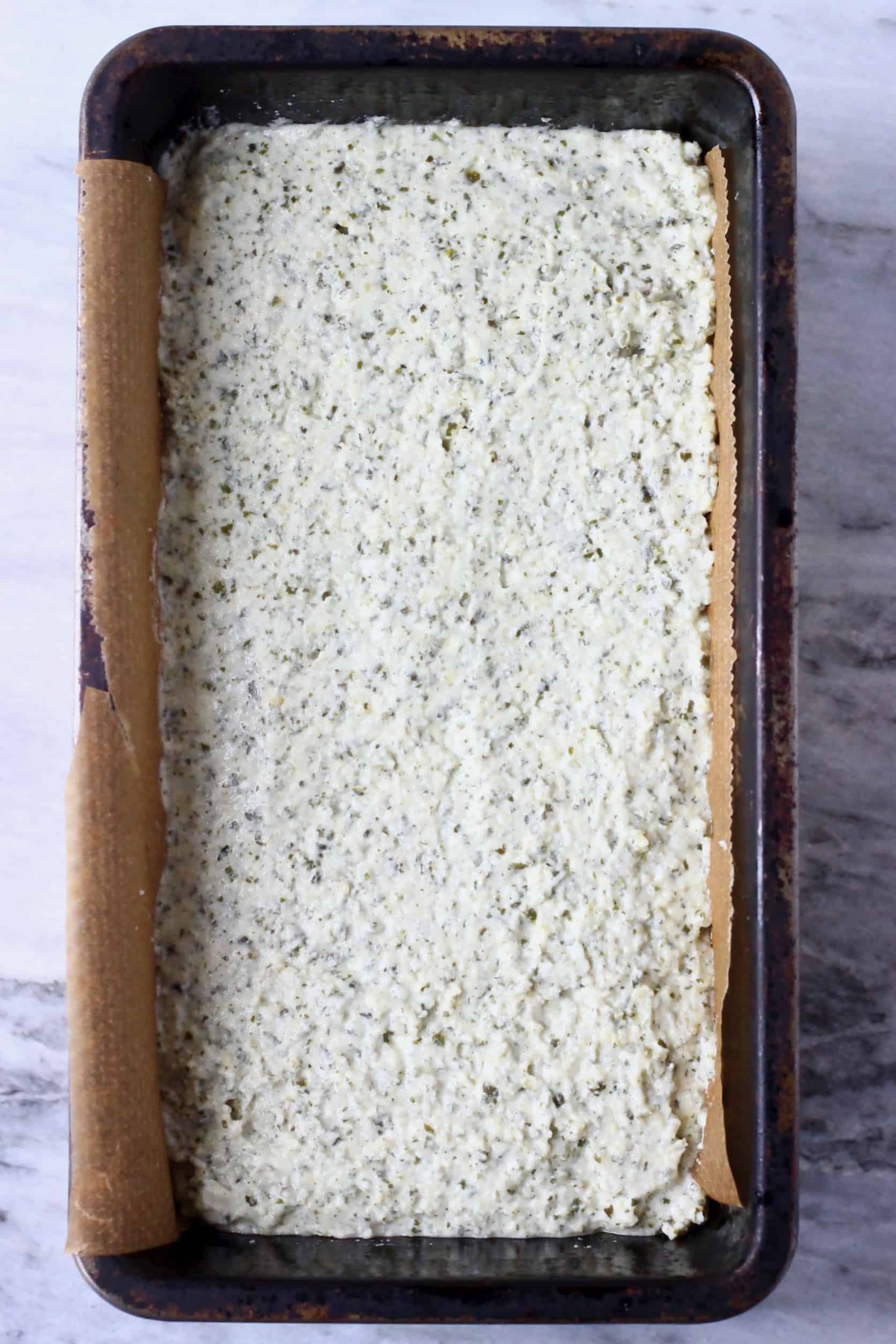 Pumpkin seed bread batter in a black loaf tin lined with baking paper against a marble background