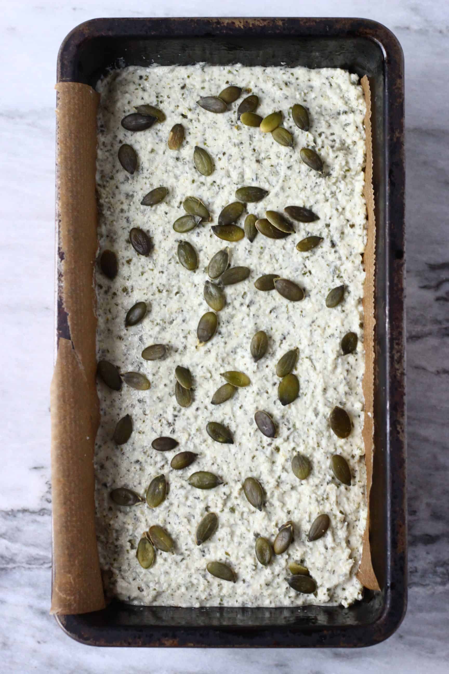 Pumpkin seed bread batter topped with pumpkin seeds in a black loaf tin lined with baking paper against a marble background