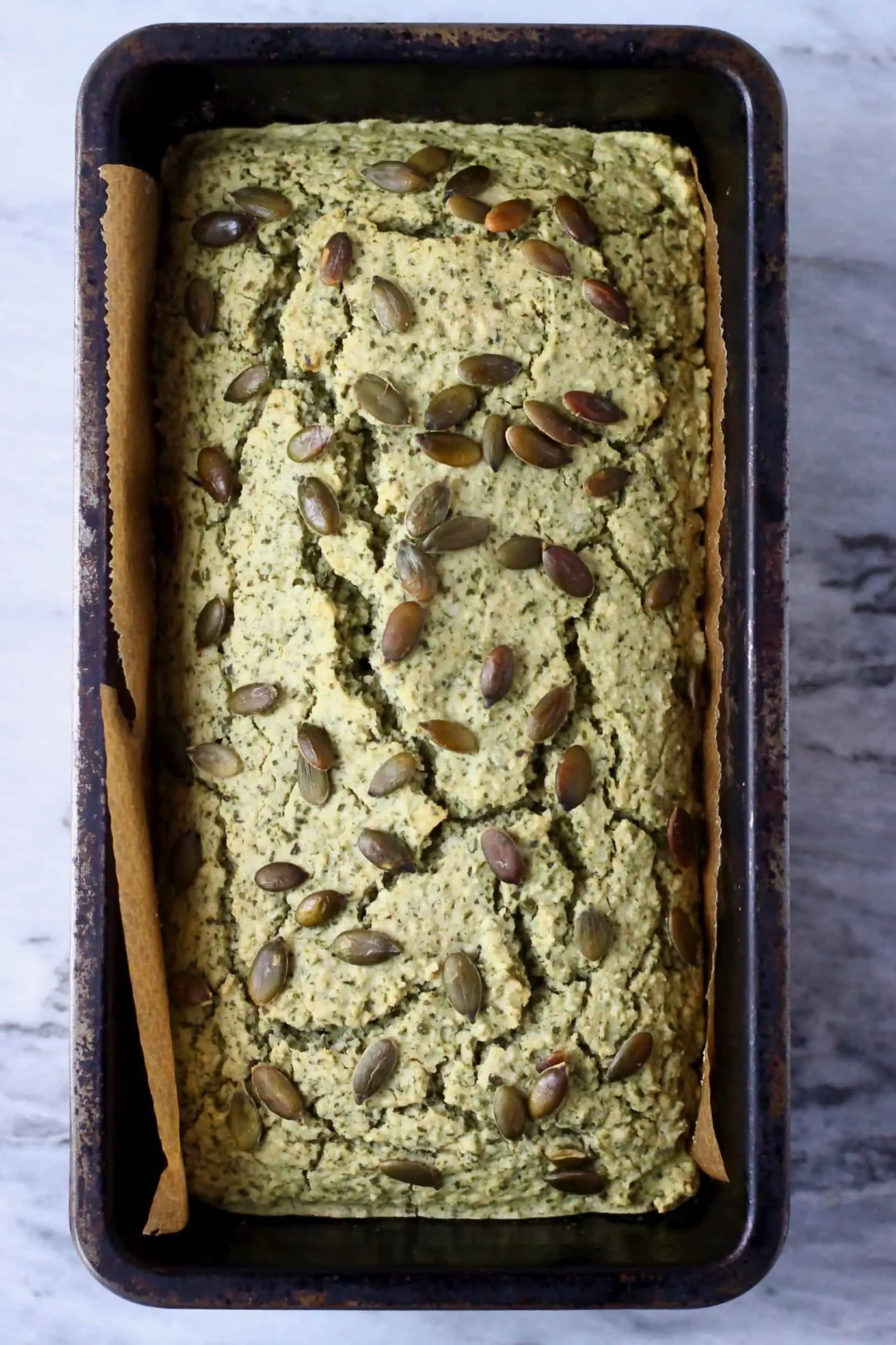 A loaf of pumpkin seed bread topped with pumpkin seeds in a black loaf tin against a marble background