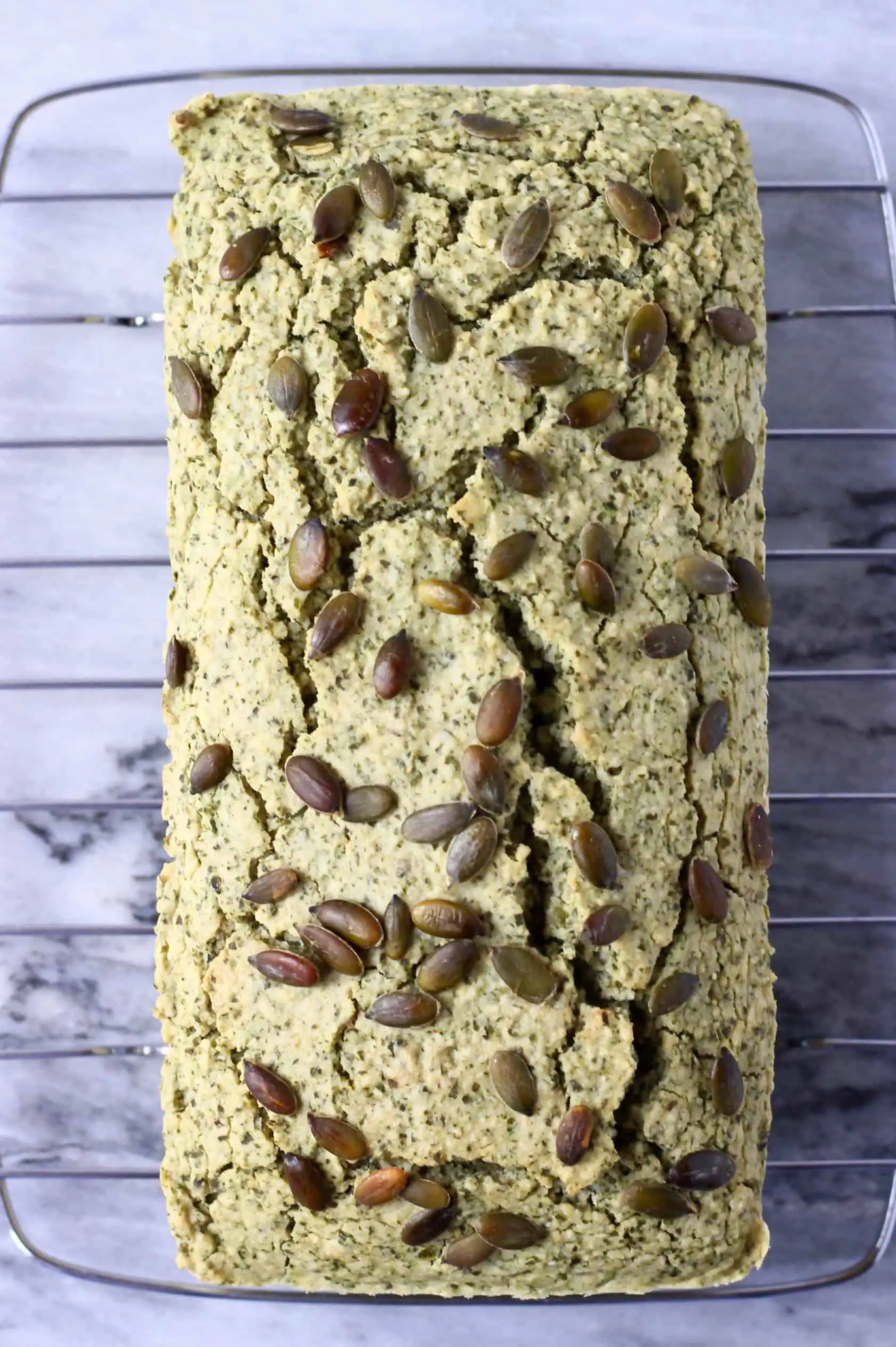 A loaf of pumpkin seed bread topped with pumpkin seeds on a wire rack against a marble background