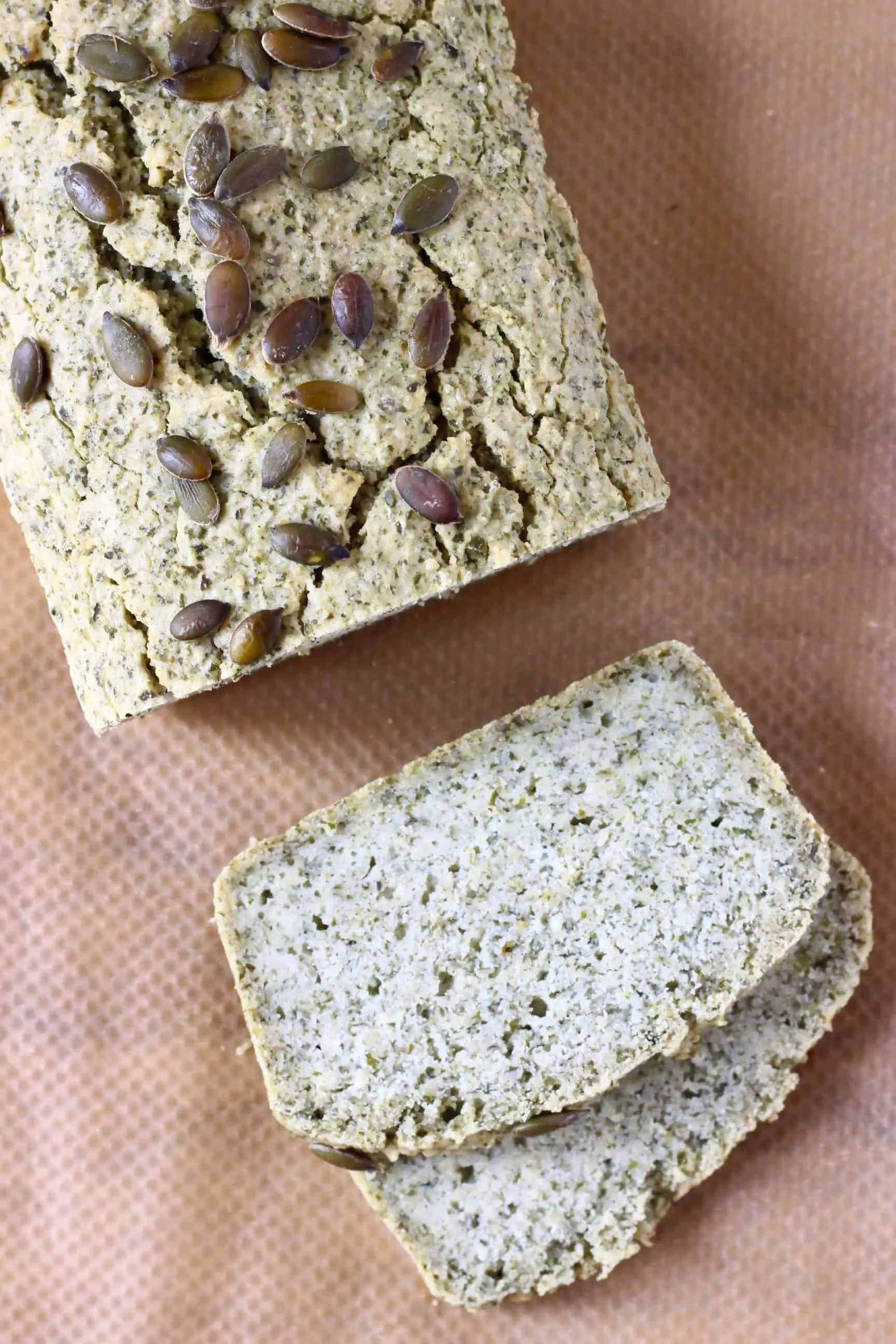 A loaf of pumpkin seed bread with two slices next to it against a sheet of brown baking paper