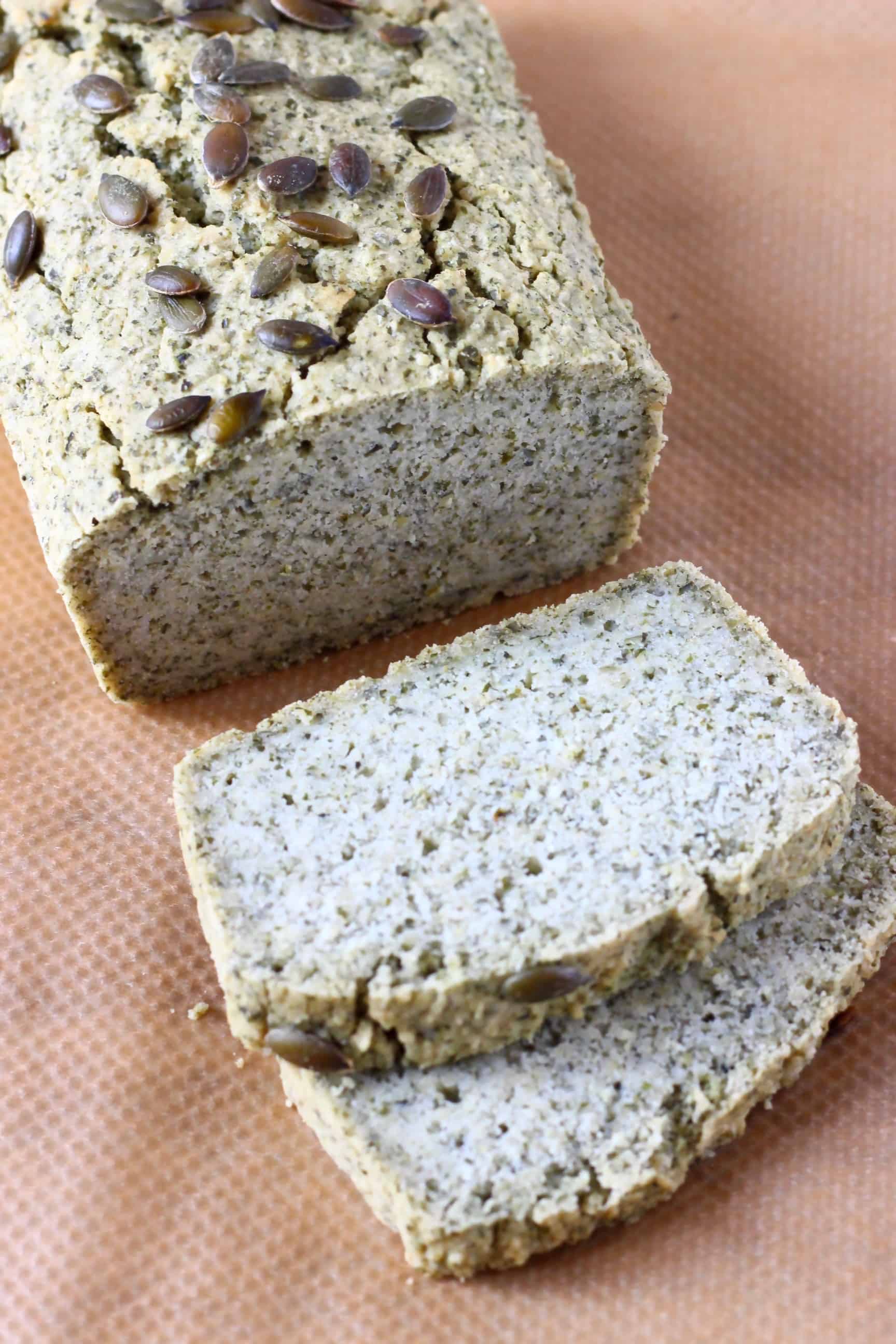 A loaf of pumpkin seed bread with two slices next to it against a sheet of brown baking paper