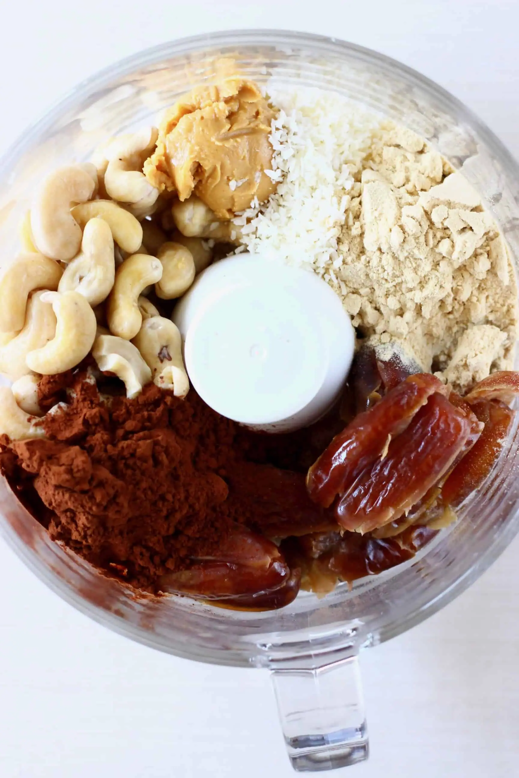 Dates, cashews, almond buter, desiccated coconut, cocoa powder and protein powder in a food processor against a white background