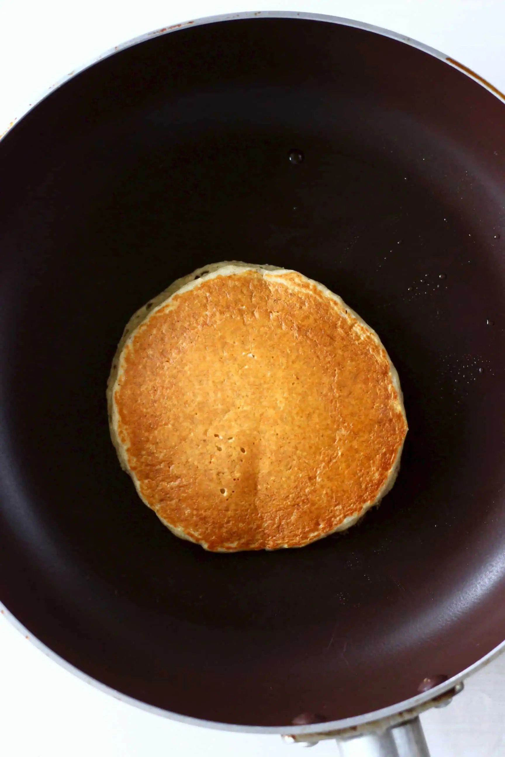 A golden brown oat flour pancake being cooked in a black frying pan against a marble background