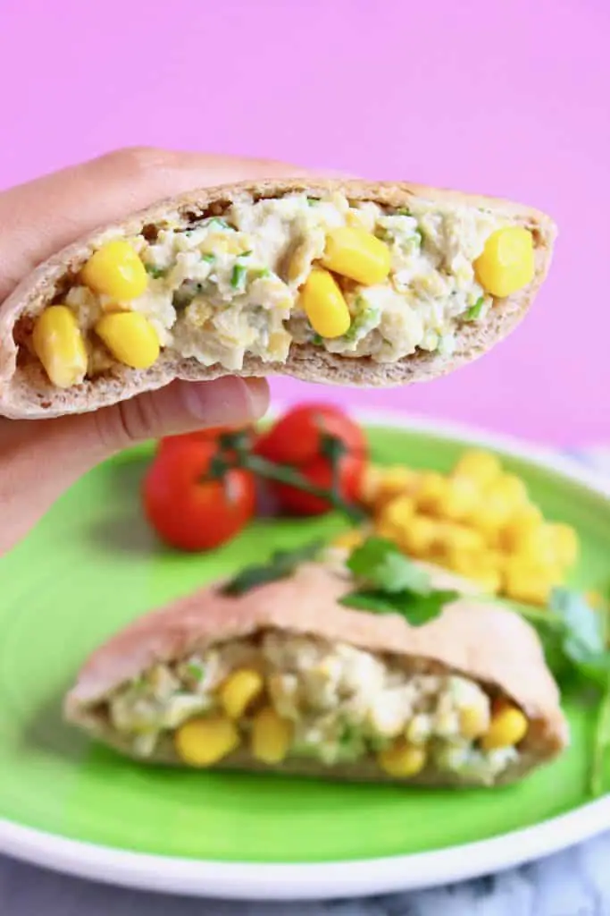 Mashed chickpeas with sweetcorn and chopped chives in mayonnaise inside a pitta on a green plate against a pink background