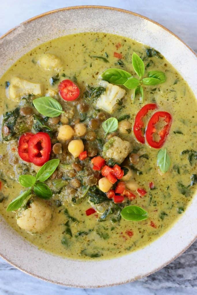 Green curry with cauliflower, spinach, chickpeas and green lentils topped with basil and red chilli in a beige bowl against a marble background