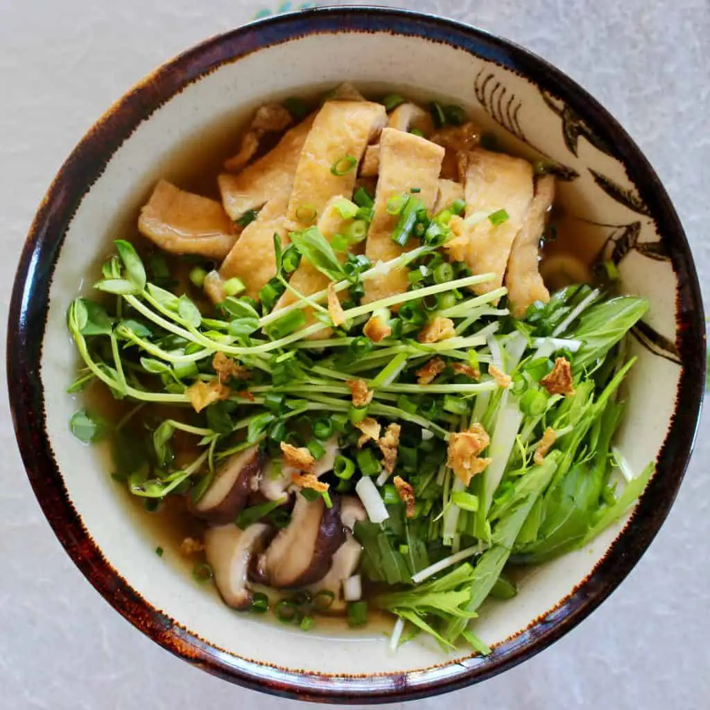 Buckwheat noodle soup topped with pieces of tofu, shiitake mushrooms and greens in a light brown bowl with a dark brown rim against a white background